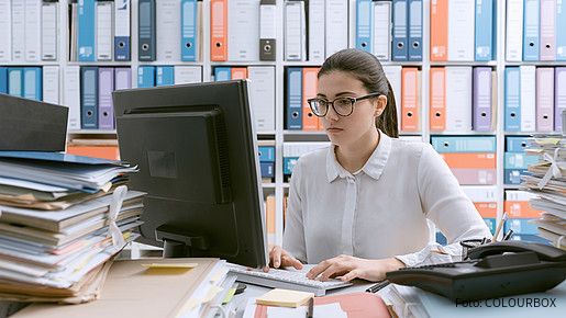 Junge Behördenmitarbeiterin sitzt am Schreibtisch hinter einem Berg von Akten, auch im Hintergrund stapeln sich Akten im Regal. 