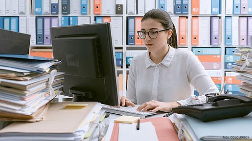 Junge Behördenmitarbeiterin sitzt am Schreibtisch hinter einem Berg von Akten, auch im Hintergrund stapeln sich Akten im Regal. 