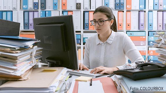 Junge Behördenmitarbeiterin sitzt am Schreibtisch hinter einem Berg von Akten, auch im Hintergrund stapeln sich Akten im Regal. 