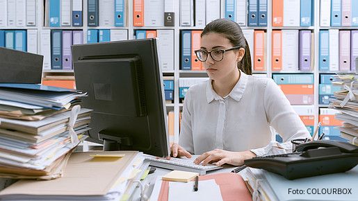 Junge Behördenmitarbeiterin sitzt am Schreibtisch hinter einem Berg von Akten, auch im Hintergrund stapeln sich Akten im Regal. 