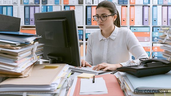 Junge Behördenmitarbeiterin sitzt am Schreibtisch hinter einem Berg von Akten, auch im Hintergrund stapeln sich Akten im Regal. 