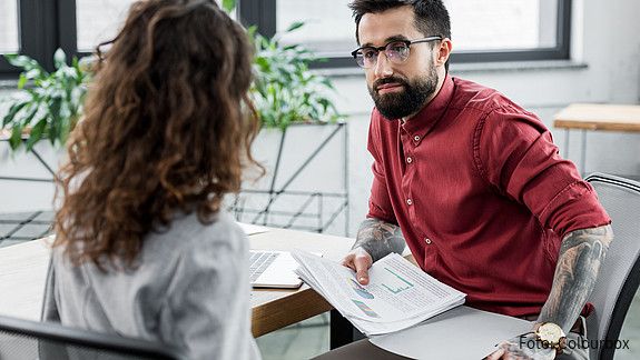 Eine von hinten zu sehende Frau und ein Mann mit tätowierten Unterarmen sitzen sich in einer Bürosituation gegenüber und sprechen miteinander.