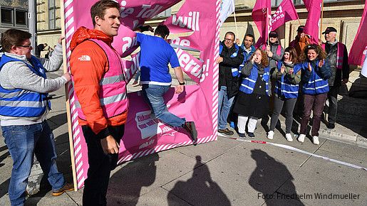 Mann springt durch ein Papier-Plakat. Um ihn herum stehen zahlreiche Demonstrierende und feuern ihn an.