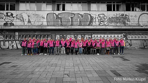 Eine Gruppe junger Beschäftigter des öffentlichen Dienstes in Warnwesten steht vor einem abrissreifen Plattenbau