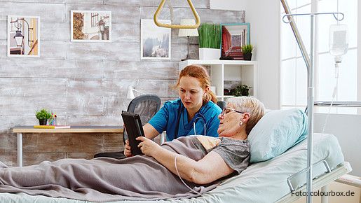 Eine ältere Frau liegt im Bett und eine Pflegerin hilft ihr dabei, ein Tablet zu bedienen